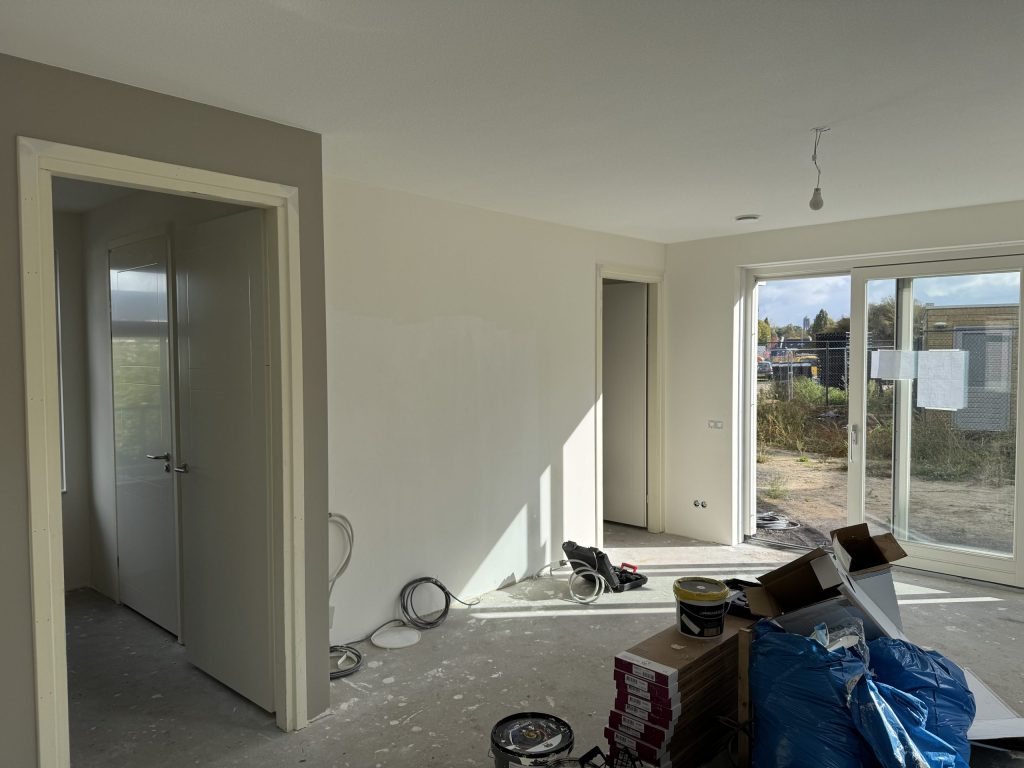 Nieuwbouwwoonkamer tijdens de afwerking, met verfemmers en materiaal op de vloer en schuifpui naar de tuin