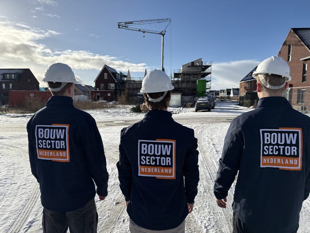 Drie bouwvakkers van Bouwsector Nederland met witte helm kijken naar een rij nieuwbouwwoningen in aanbouw onder een blauwe winterlucht